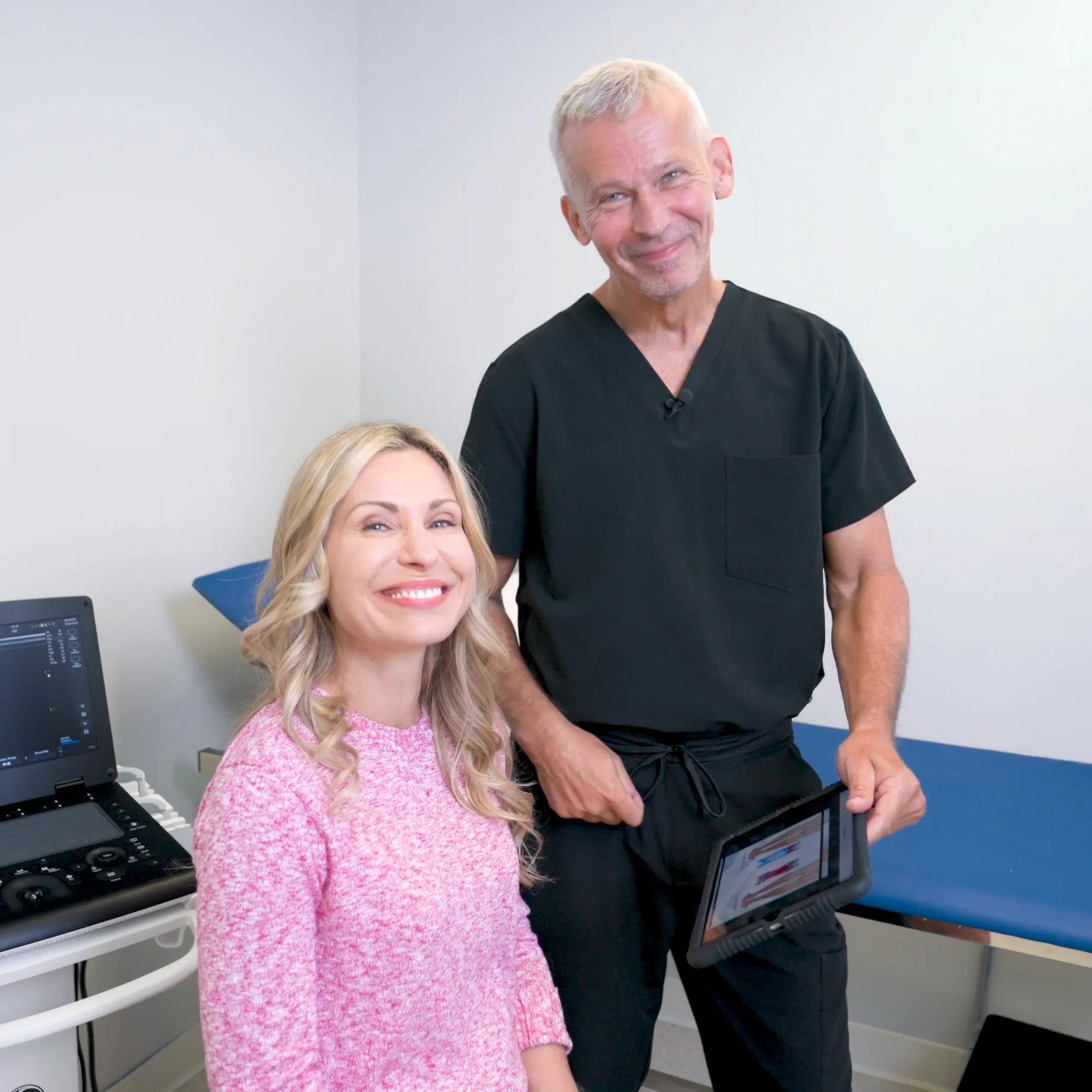 Dr. Martin Tyson stands with a patient in a clinical examination room, both smiling at the camera. He holds a tablet displaying medical imagery, with an ultrasound machine positioned nearby.